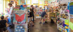   Image of toy store filled with unique hard to find toys and filled with happy playing children. In the foreground a father enjoys building toys with his son.  
