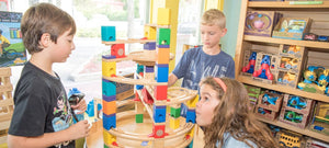   Three children look in wonder as they play with a wooden Hape marble run in a unique toy store. A shelf in the back is filled with toddler toys. 